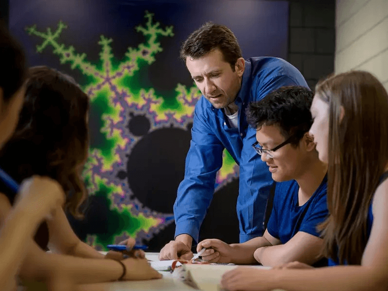 Group of students gathered around a table, one student pointing at a paper while explaining a concept.
