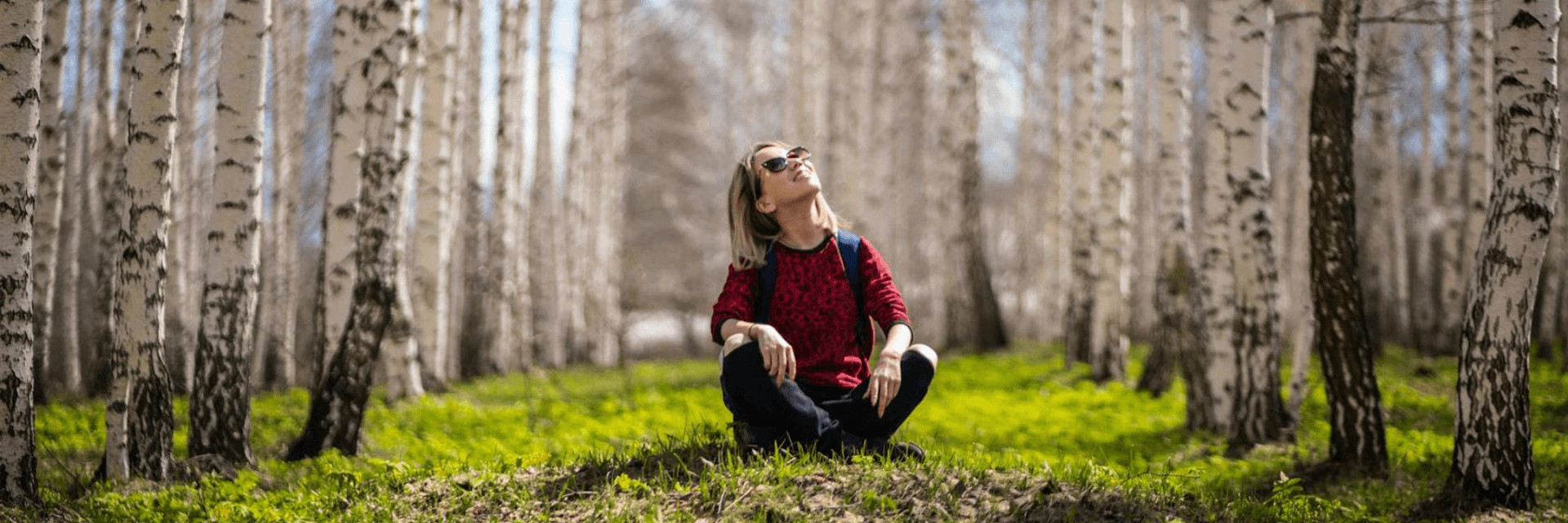 Women sitting cross legged on grass surrounded by birch trees. She is looking up at the sky